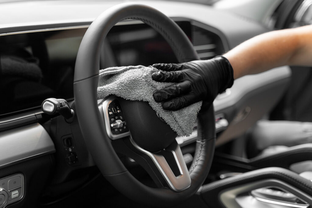 Hand in schwarzem Handschuh reinigt das Lenkrad eines Autos mit einem Mikrofasertuch.
